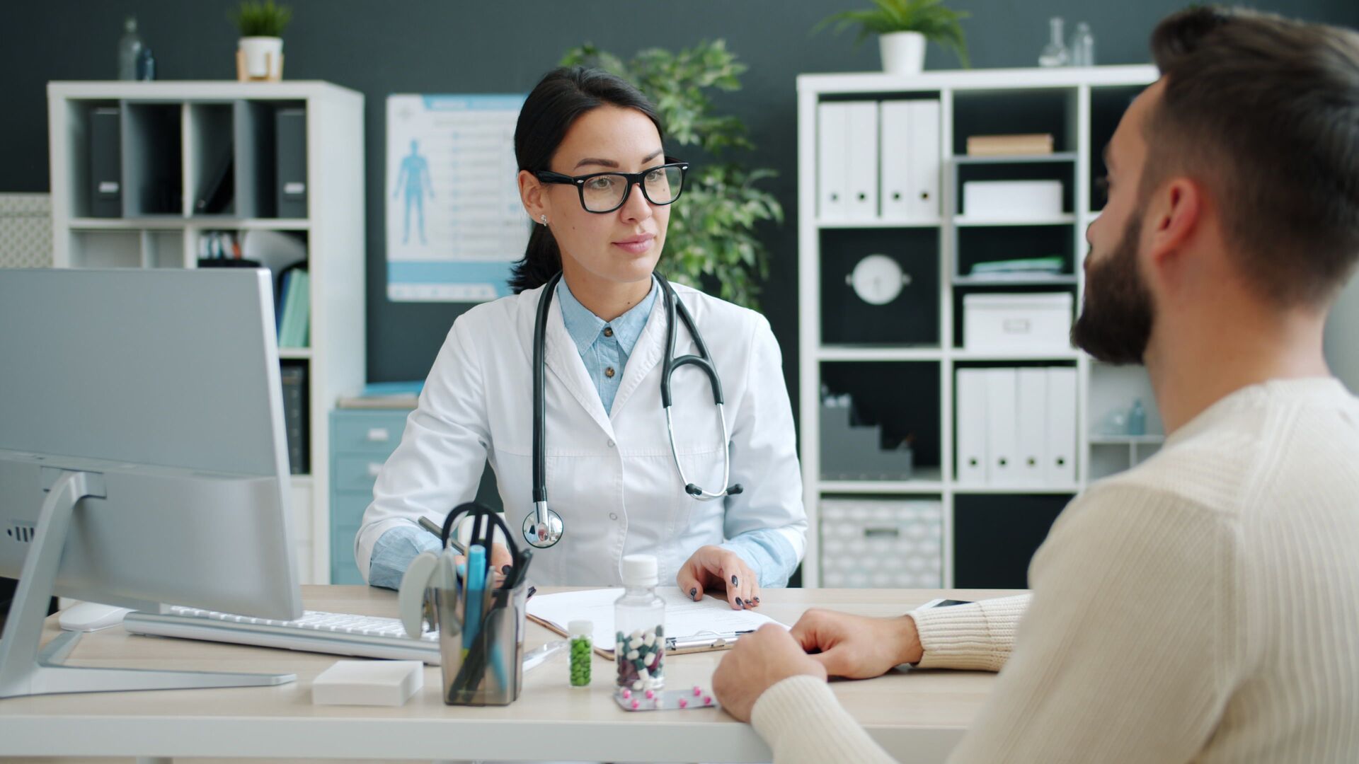 Doctor in a white coat talking with a male patient across a desk in a bright clinic office